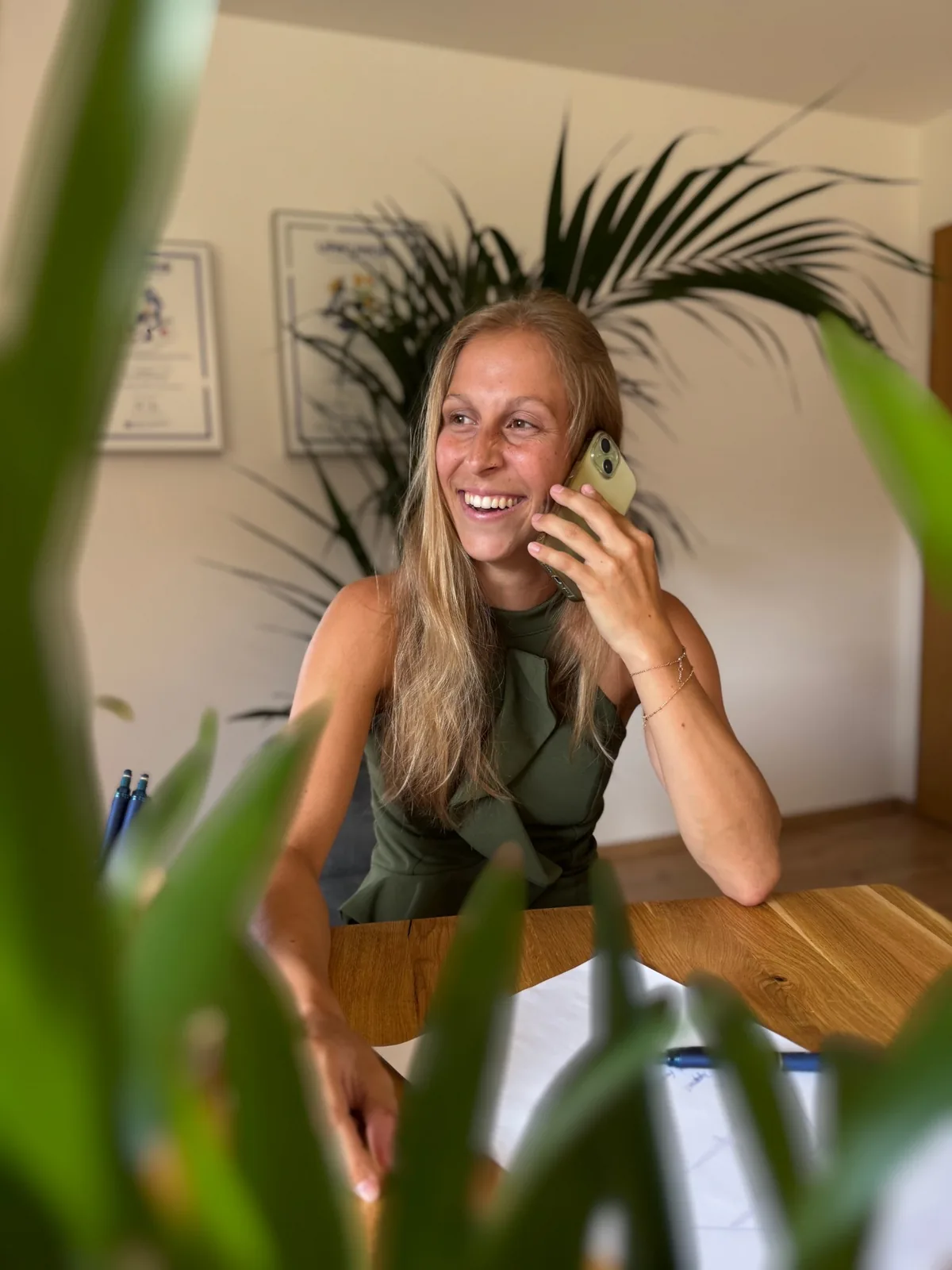 Lena Kottbauer telefoniert lächelnd an ihrem Besprechungstisch, im Hintergrund eine Zimmerpalme