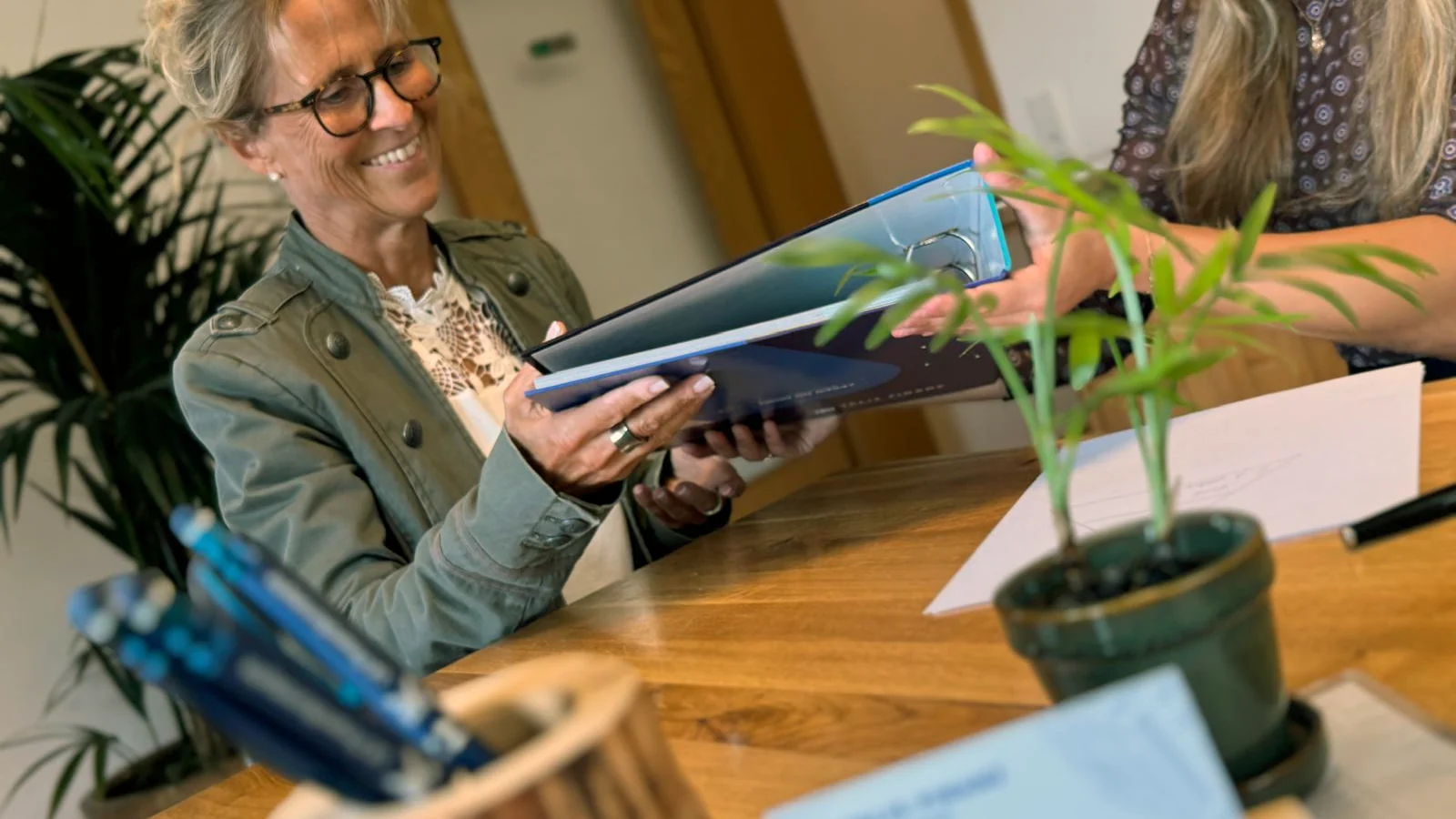 Lena reicht einer Kundin eine Unterlagenmappe über den Besprechungstisch